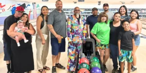 A group of smiling ARCA members and supporters stand together at the bowling alley, some holding bowling balls and others wearing event T-shirts, with colorful lanes and tournament signage in the background—capturing the spirit of community, fun, and heartfelt generosity that defines the annual charity event.